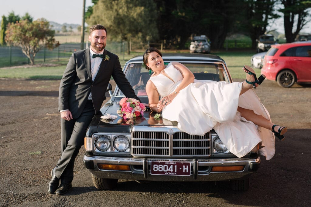 Relaxed couple sit on classic car at Warrnambool wedding.